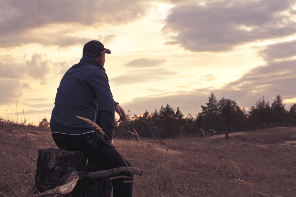 A man in a jacket sits on a stump outdoors, contemplating the serene sunset scenery.