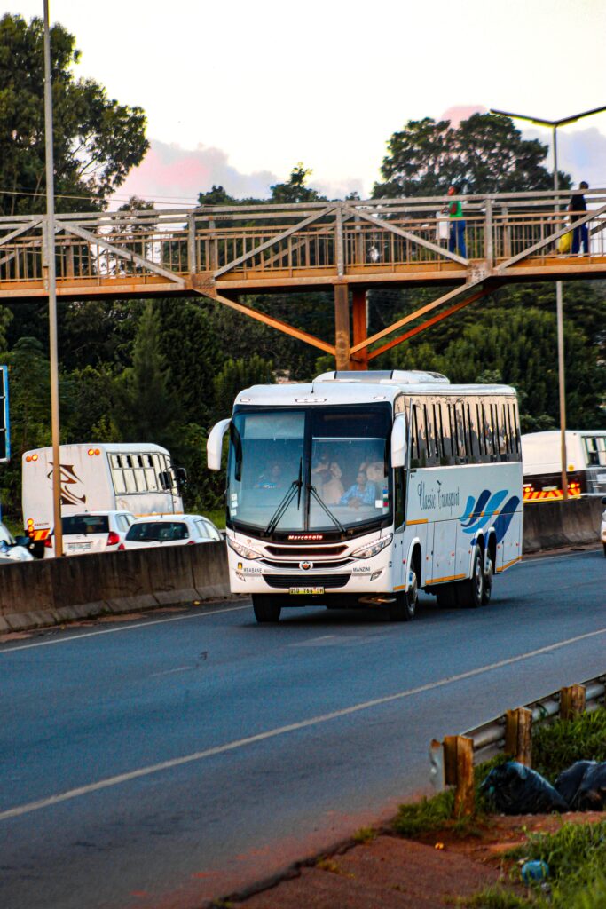 A white passenger bus travels on a highway beneath an overpass in Matsapha, Eswatini during the day.